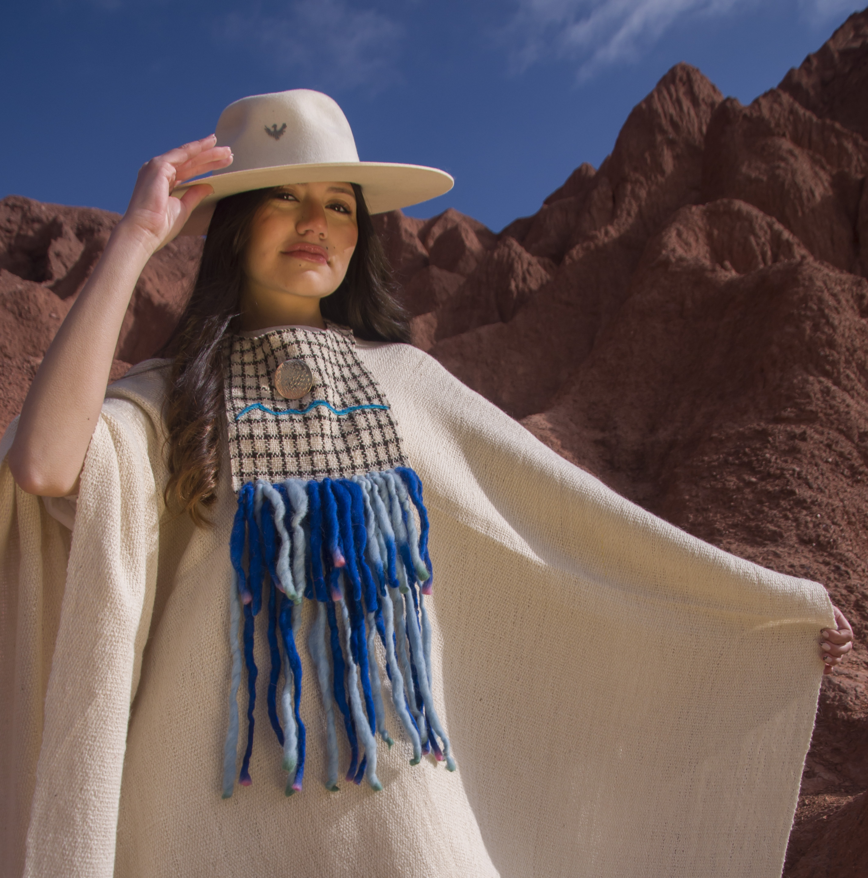 Mujer con poncho de fibra de llama color crudo, sombrero blanco y flecos azules, con los brazos extendidos en la Quebrada de Purmamarca, Jujuy