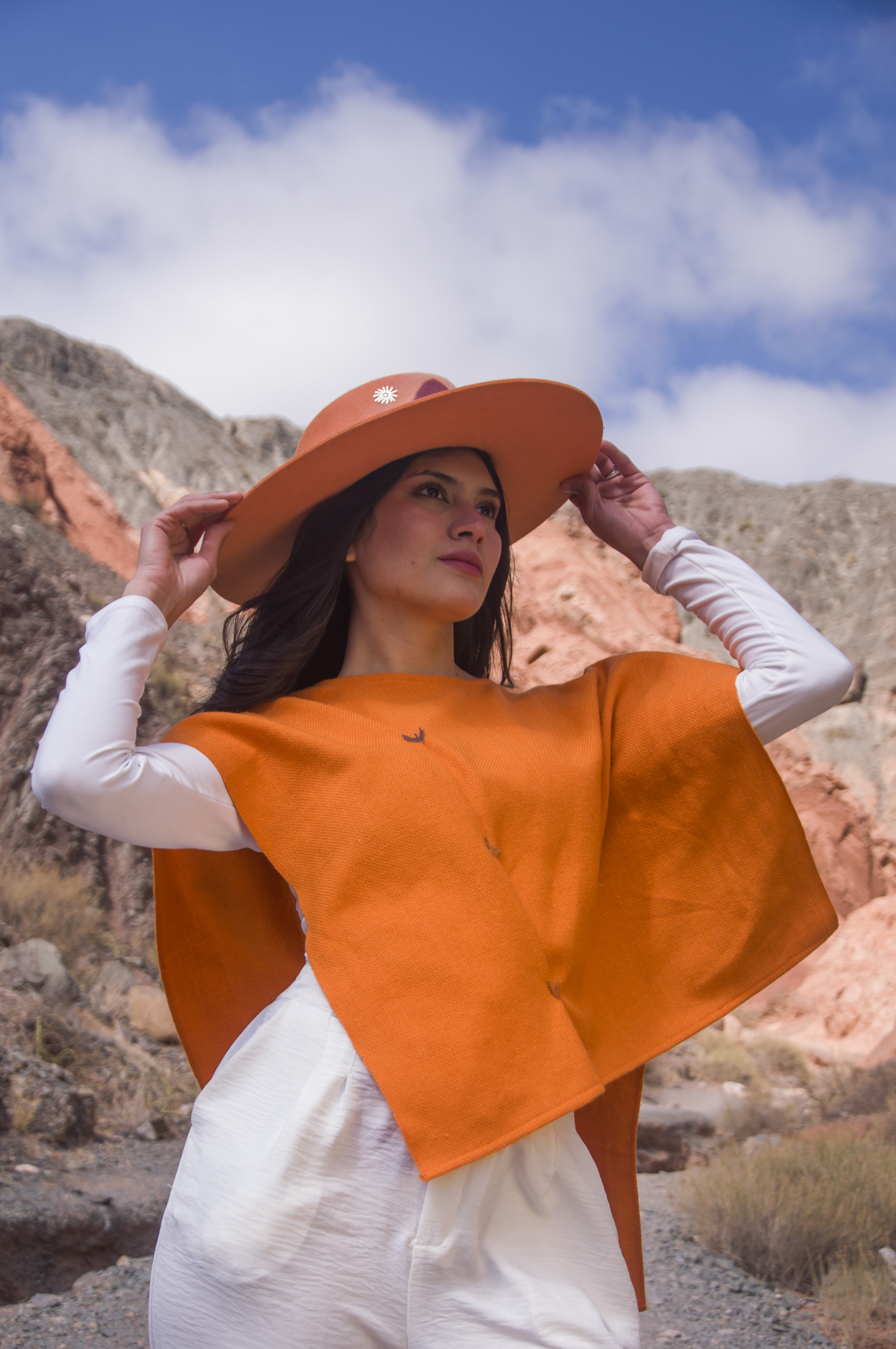 Mujer con top de fibra de llama color naranja y sombrero a tono, entre los cerros de colores de Purmamarca